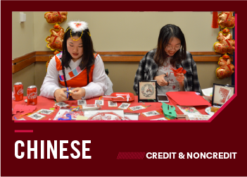 Two women sit at a table making paper cutouts surrounded by festive decorations.