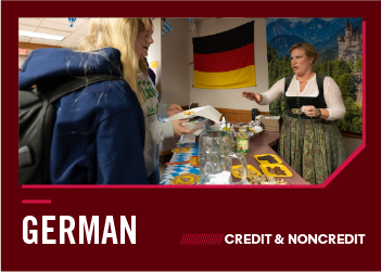 A woman in a traditional dirndl offers food to students in front of a German flag.